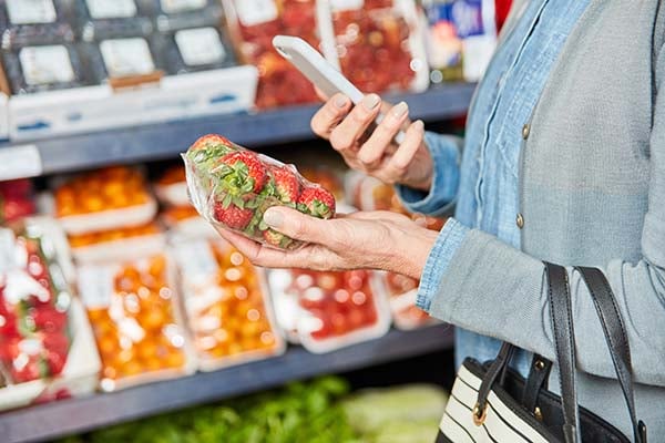 Shopper holding a pack of strawberries and a smartphone in a grocery store produce aisle