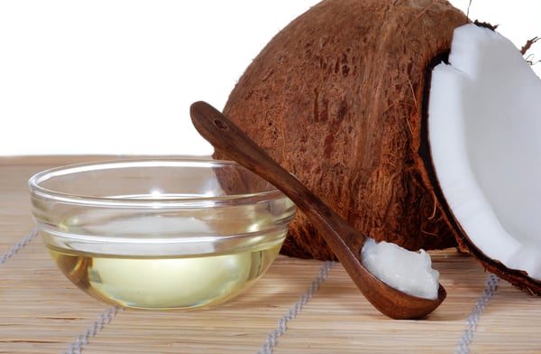 Glass bowl of coconut oil and a wooden spoon of solid coconut oil beside a halved coconut