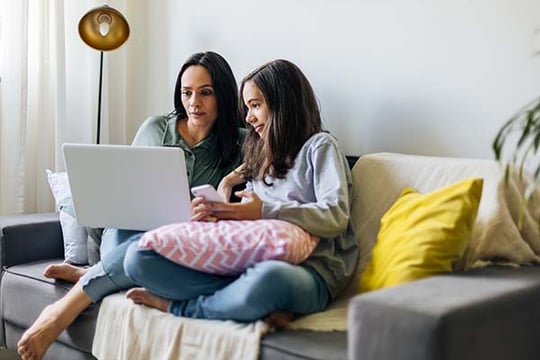 Woman and teen girl sitting together on a couch, looking at a laptop and a smartphone