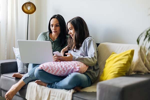 Woman and teen girl sitting together on a couch, looking at a laptop and a smartphone