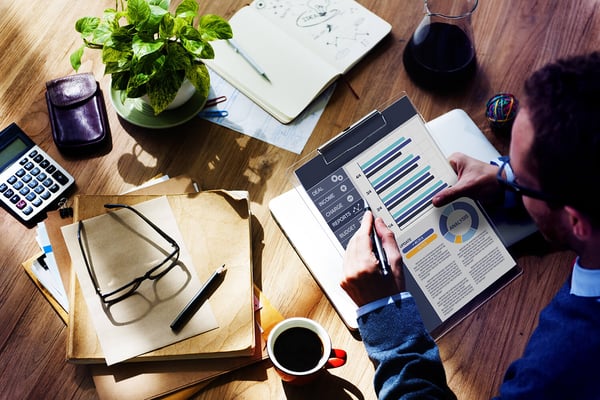 Man reviewing financial charts on a clipboard at a desk with a calculator, notebook and coffee