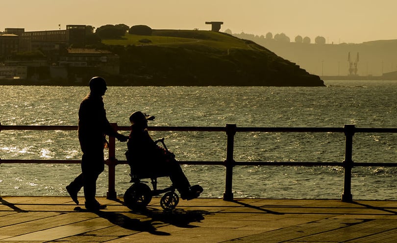Silhouette of a person pushing someone in a wheelchair along a waterfront walkway at sunset