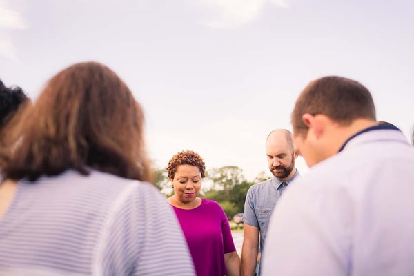 Small group of adults standing in a circle outdoors with heads bowed in prayer