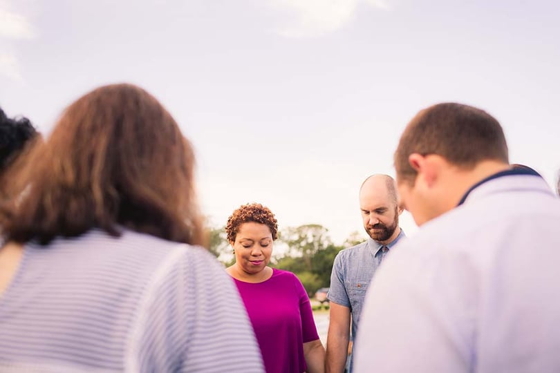 Small group of adults standing in a circle outdoors with heads bowed in prayer