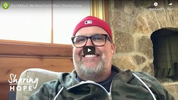 Bart Millard in a red cap smiling by a stone fireplace in a still from the Sharing Hope video We Need Each Other