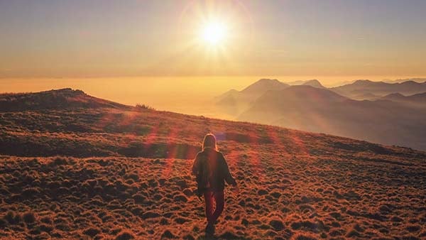 Lone hiker walking across a grassy mountain ridge toward a bright sun over distant peaks