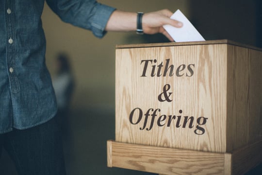 Person dropping an envelope into a wooden box labeled Tithes & Offering