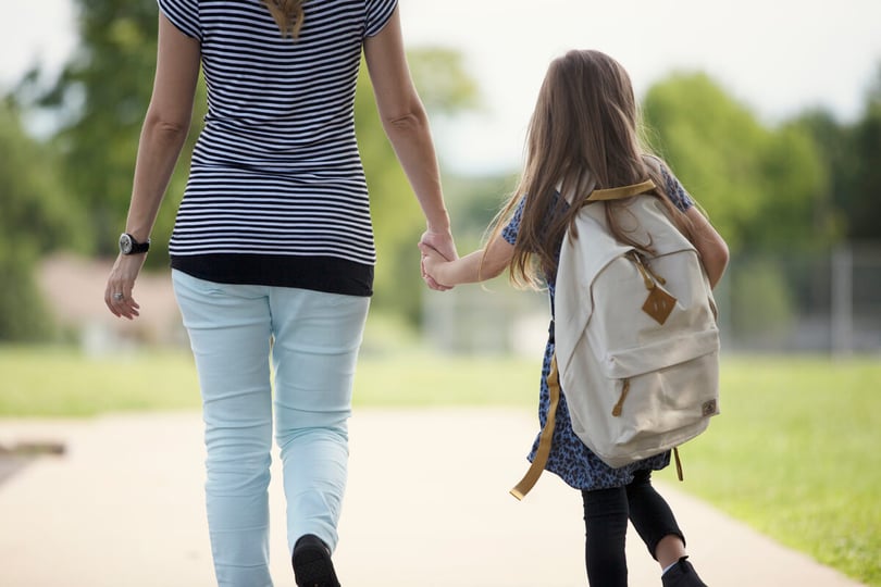 Woman and a young girl with a backpack walk hand in hand along a path, seen from behind