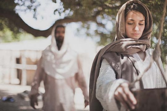 Biblical-era scene of a woman drawing water at a well as a man in white robes approaches behind her