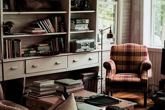 Cozy reading room with crowded bookshelves, a plaid armchair by the window and books stacked on a table