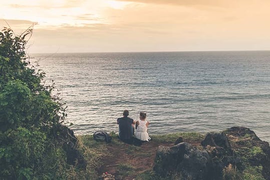 Two people sitting on a grassy clifftop looking out over the ocean at sunset