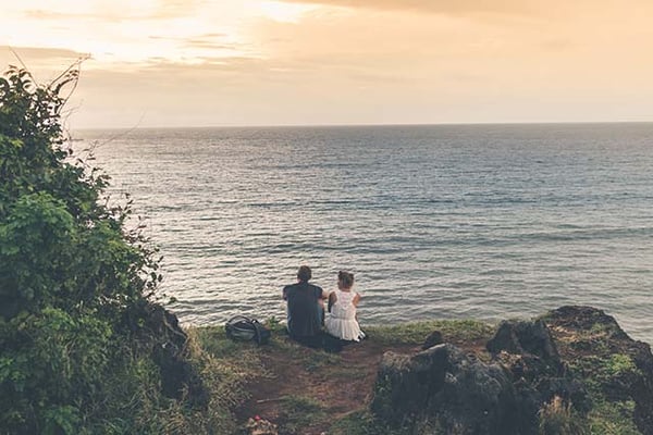 Two people sitting on a grassy clifftop looking out over the ocean at sunset