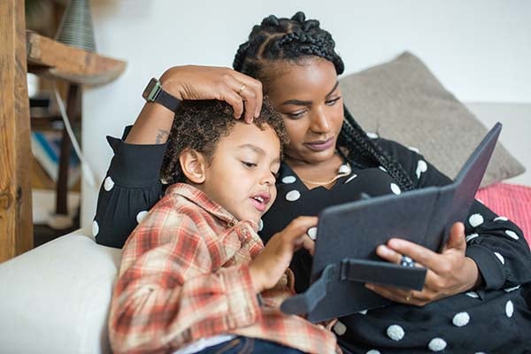 Woman and a young boy reading together on a tablet while relaxing on a couch