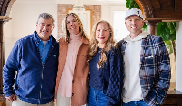 Four smiling adults standing together in a bright home, one man wearing a cap with a cross