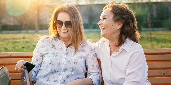 Middle-aged woman and older woman laughing together on a park bench in golden sunlight