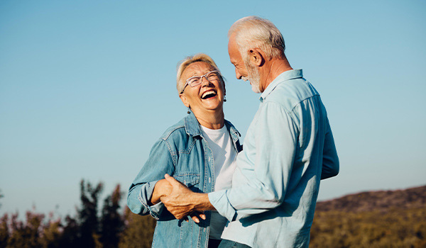 Older man and woman laughing as they dance outdoors under a clear blue sky