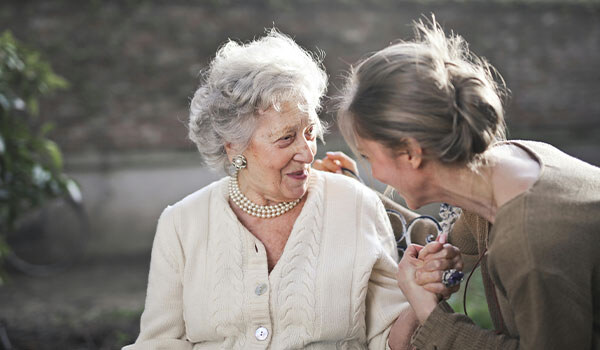 Smiling older woman in a cardigan and pearls holds hands with a younger woman as they talk outdoors
