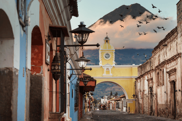 Yellow Santa Catalina Arch over a cobblestone street in Antigua, Guatemala, with a volcano rising behind it