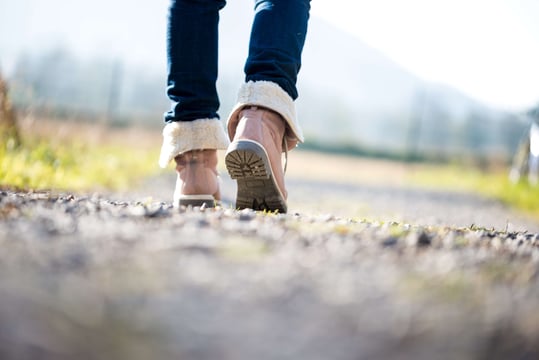 Low-angle view of a person in fleece-lined boots and jeans walking along a sunlit gravel path