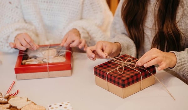 Two people in cozy sweaters tying twine bows on holiday gift boxes, with cookies and candy canes nearby
