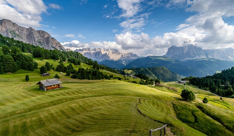 Green alpine meadows with a wooden hut and pine forests below jagged rocky peaks under a bright, cloudy sky