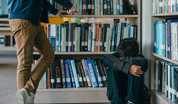 Teen sitting hunched on a library floor with head down as another person stands over them, pointing