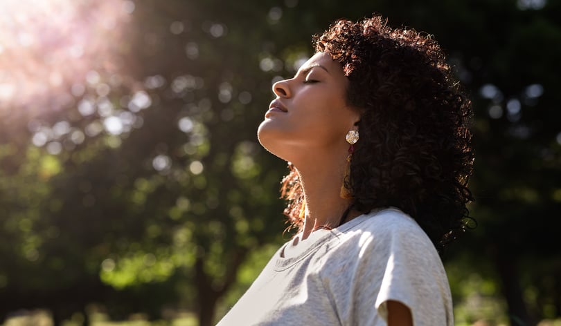 Woman with eyes closed tilting her face toward the sun outdoors among green trees
