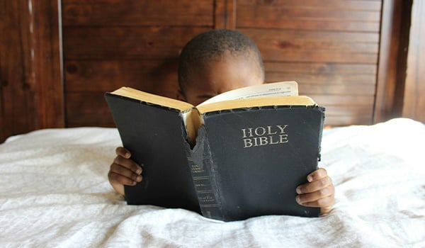 Young child on a bed reading a large, worn Holy Bible held up close to their face