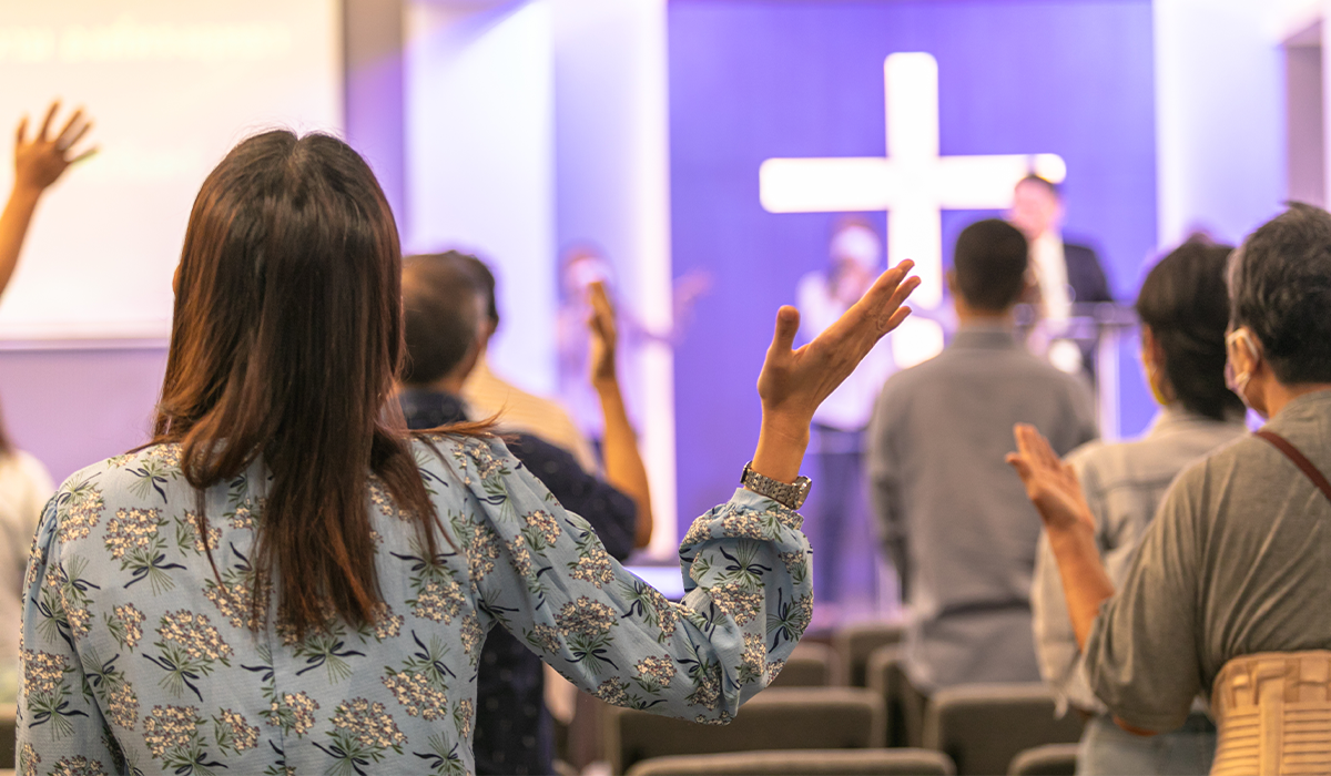 People participate in a worship service, raising hands in a church sanctuary with a cross at the front.