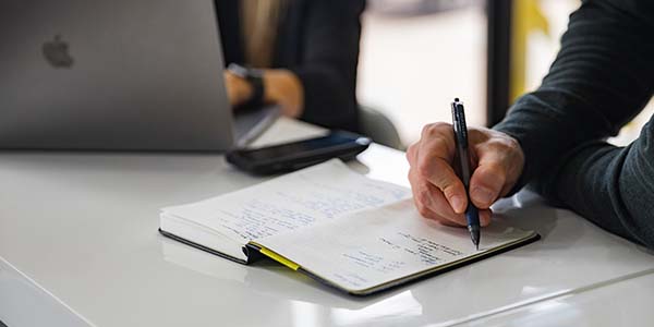 close up of hand writing notes sitting next to person at computer