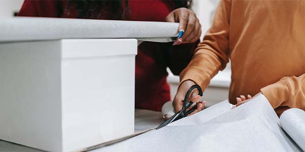 Close-up of two people wrapping a white gift box, one cutting the paper with scissors