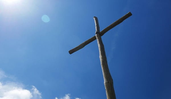 Rough wooden cross seen from below against a deep blue sky with sunlight and a few white clouds