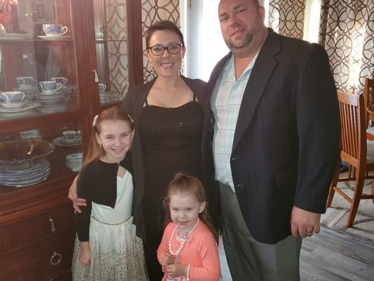 Smiling family of four, two adults and two young girls, posing in a dining room beside a china cabinet