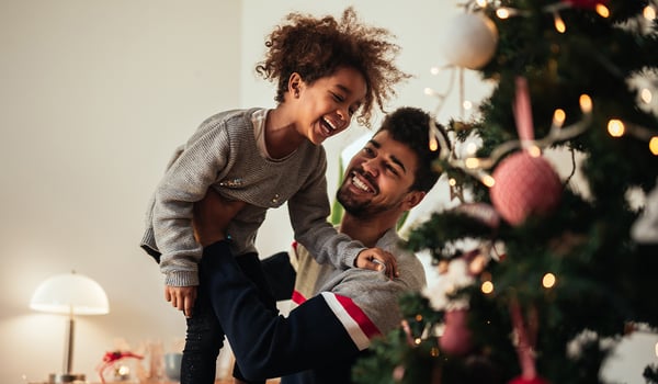 Smiling man lifting a laughing young girl into the air beside a lit Christmas tree