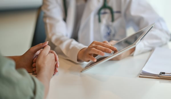 Doctor in a white coat with a stethoscope shows a tablet to a patient across a desk