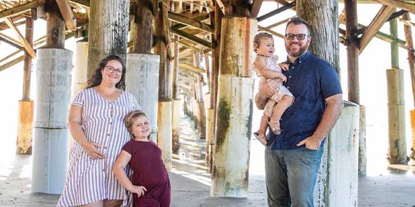 Dom Robin and his family standing together beneath a wooden beach pier