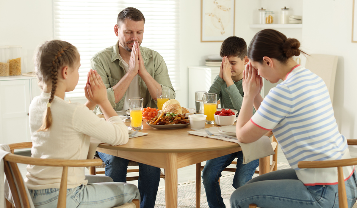 People sit around a dining table with hands held together before a meal, with food and drinks on the table.