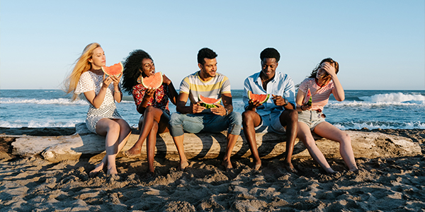 Five young adults sitting on a driftwood log at the beach, eating slices of watermelon