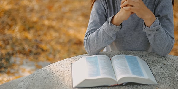 Person in a gray sweater with hands clasped in prayer over an open Bible on a stone table outdoors