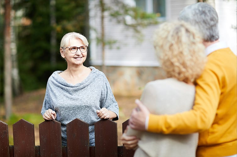 Blonde mature woman in eyeglasses and grey pullover looking at her neighbors during talk through fence