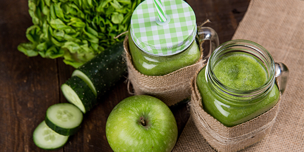 Green smoothies in mason jars with a green apple, sliced cucumber and leafy greens on a wooden table