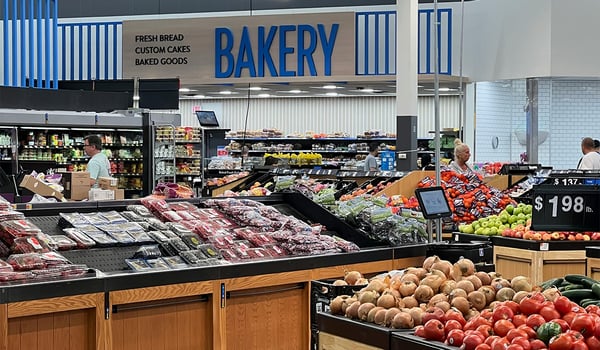Supermarket produce section with bins of tomatoes, onions, apples and berries beneath a blue Bakery sign