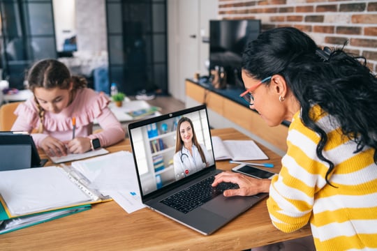 Woman on a laptop video call with a doctor while a girl does homework at the same table