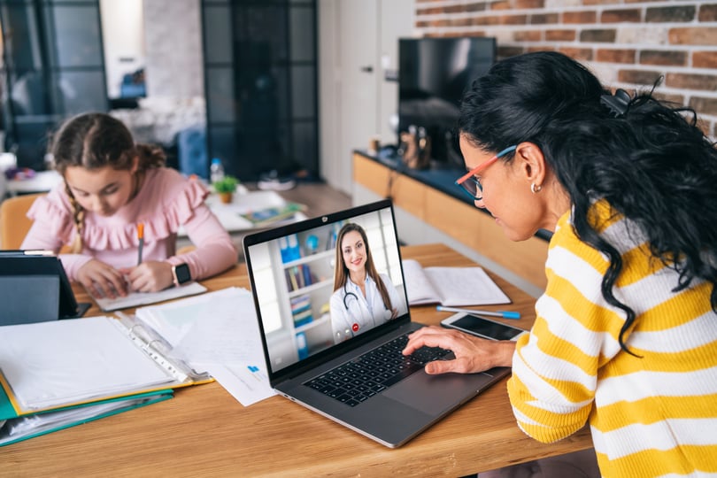 Woman on a laptop video call with a doctor while a girl does homework at the same table