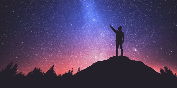 A man pointing to the stars while he stands at the top of a hill