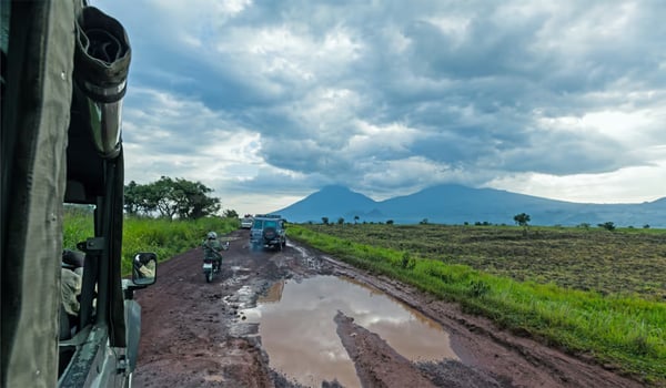 View from a vehicle down a muddy rural road, a motorbike and another vehicle ahead, mountains under storm clouds