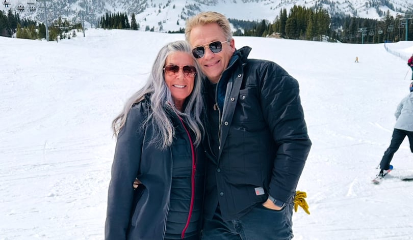 Paul and Kelli are wearing winter clothing stand on a snowy ski slope with trees and mountains behind them.
