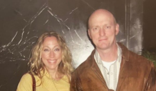 Kellye, in a yellow top, and Baron, in a brown leather jacket, smile together in front of a dark marble wall