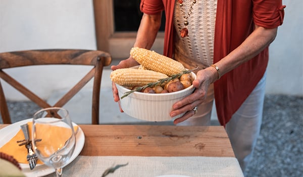 Woman carries a bowl of corn on the cob and baby potatoes to an outdoor table set for a meal