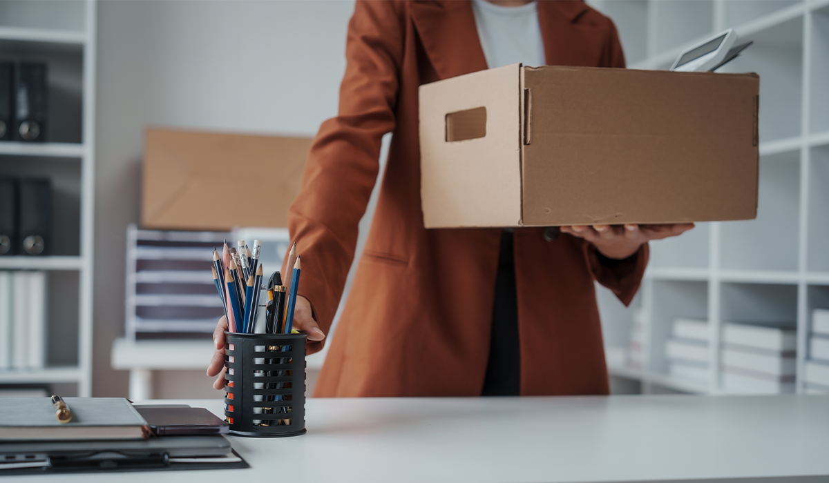 Person gathers office supplies from a desk while holding a cardboard box in an office setting.